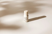 A small cream-colored bottle with a ribbed cap stands on a warm beige surface under soft, directional sunlight that casts a long shadow. The label reads 'French Orchard' and 'vörda'.