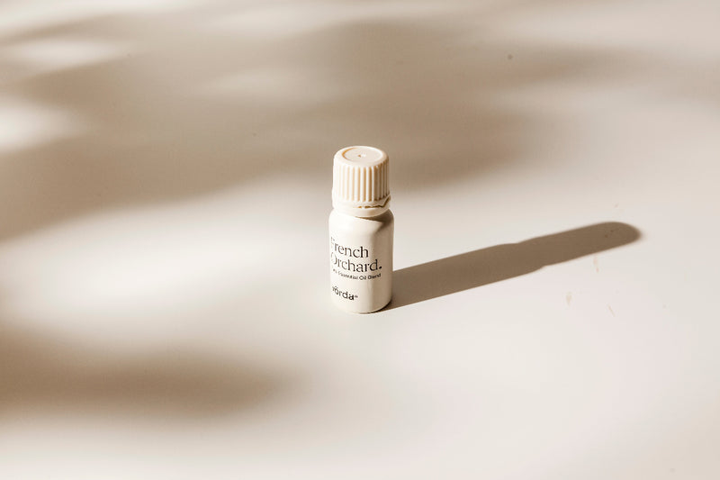 A small cream-colored bottle with a ribbed cap stands on a warm beige surface under soft, directional sunlight that casts a long shadow. The label reads 'French Orchard' and 'vörda'.