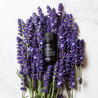 A small black bottle labeled 'Spike Lavender' and 'Vörda' is nestled in the center of a dense cluster of purple lavender flowers on a light marble-like background.