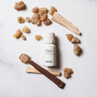 A small white bottle labeled 'Zen Pure Essential Oil' by vördä, arranged on a light marble-like surface with scattered amber cluster pieces and two palo Santo sticks.