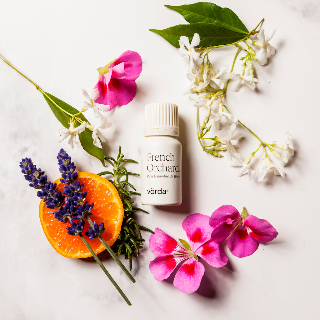 A small white bottle labeled 'French Orchard' and 'vördä' is arranged on a light marble surface with flowers, a sliced orange, lavender sprigs, and green leaves around it.