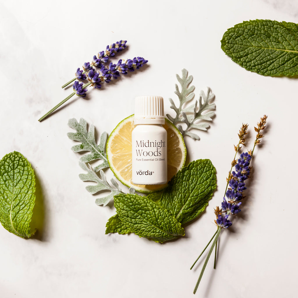 White essential oil bottle labeled 'Midnight Woods' and 'vörda' over a slice of ice, mint leaves, lavender stems, and foliage on a white background