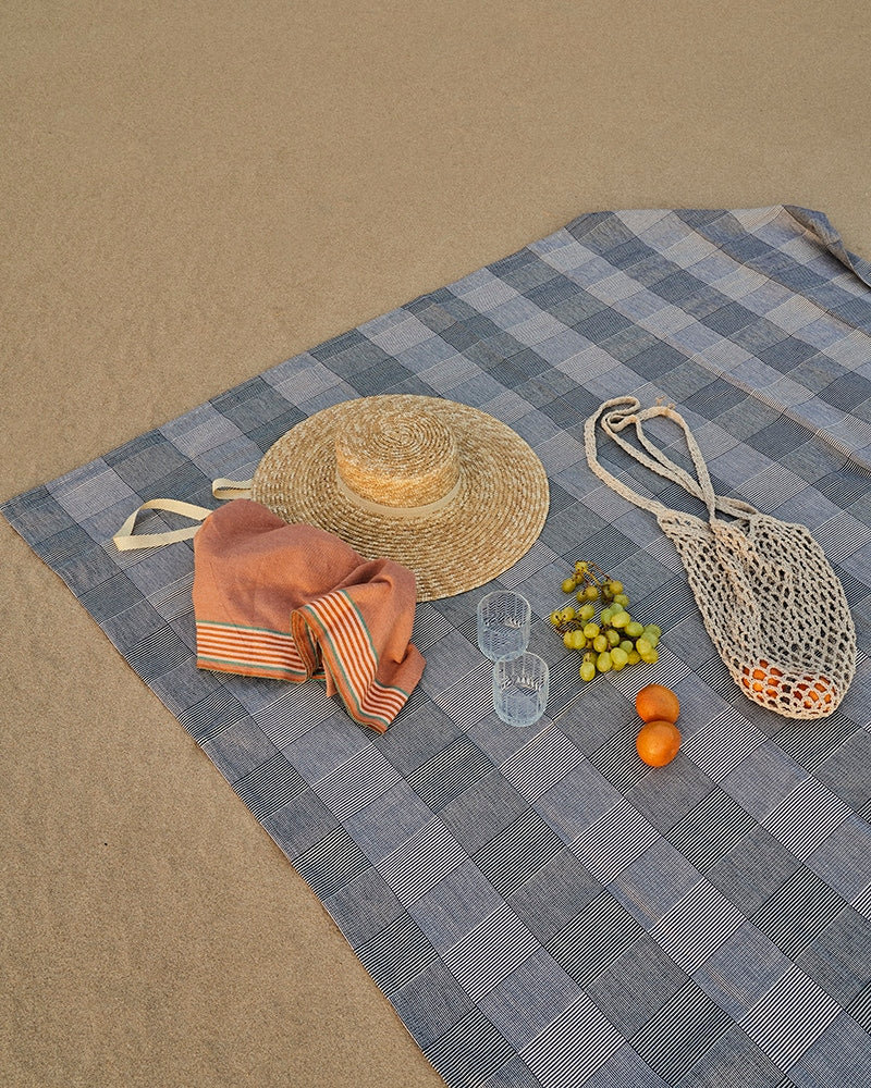 Blue checkered picnic blanket with a straw hat, orange, fruits, and a bag on the sand
