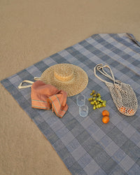 Blue checkered picnic blanket with a straw hat, orange, fruits, and a bag on the sand