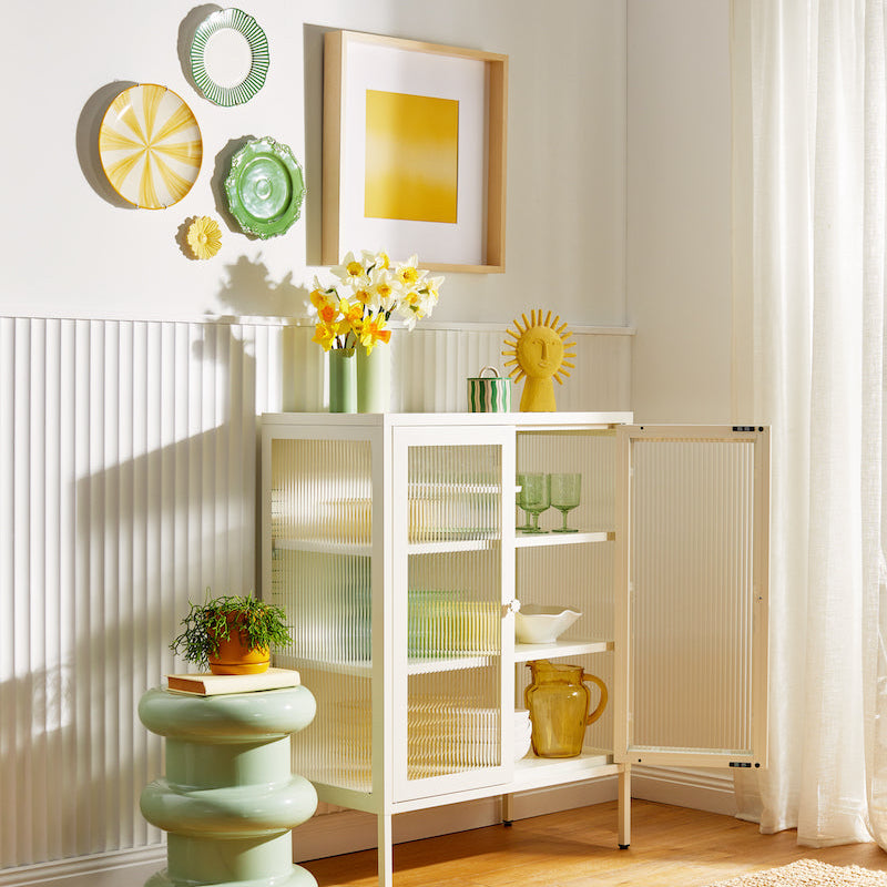 White locker with glass doors and one door ajar holding and surrounded by yellow and light green home decor in a white sunlit room