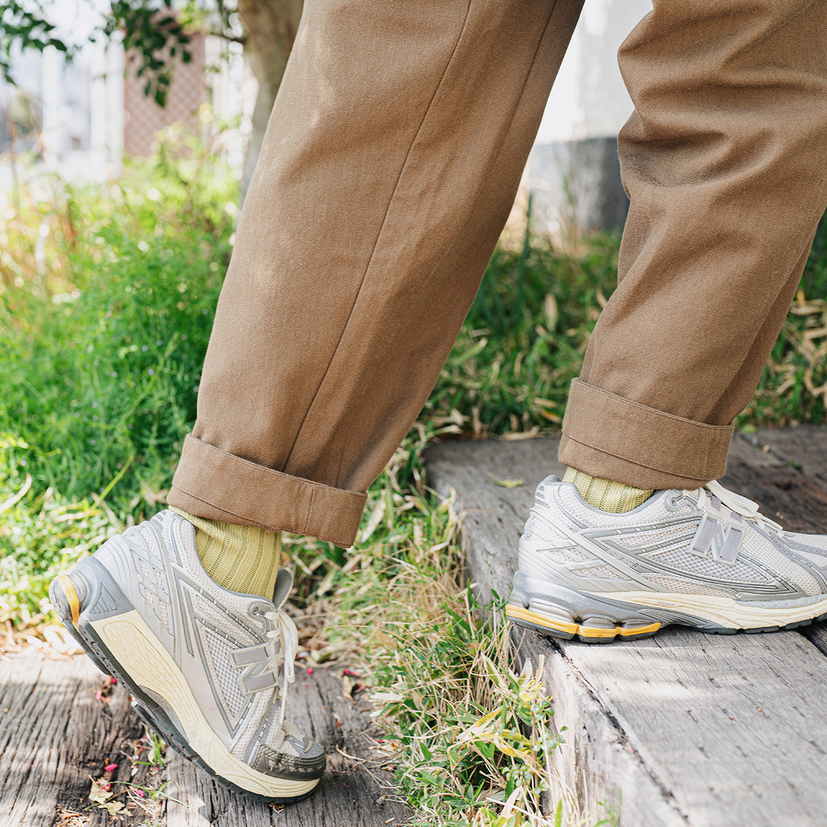 Person's legs wearing brown pants, light green socks, and grey athletic shoes, stepping on wooden beam stairs in grass