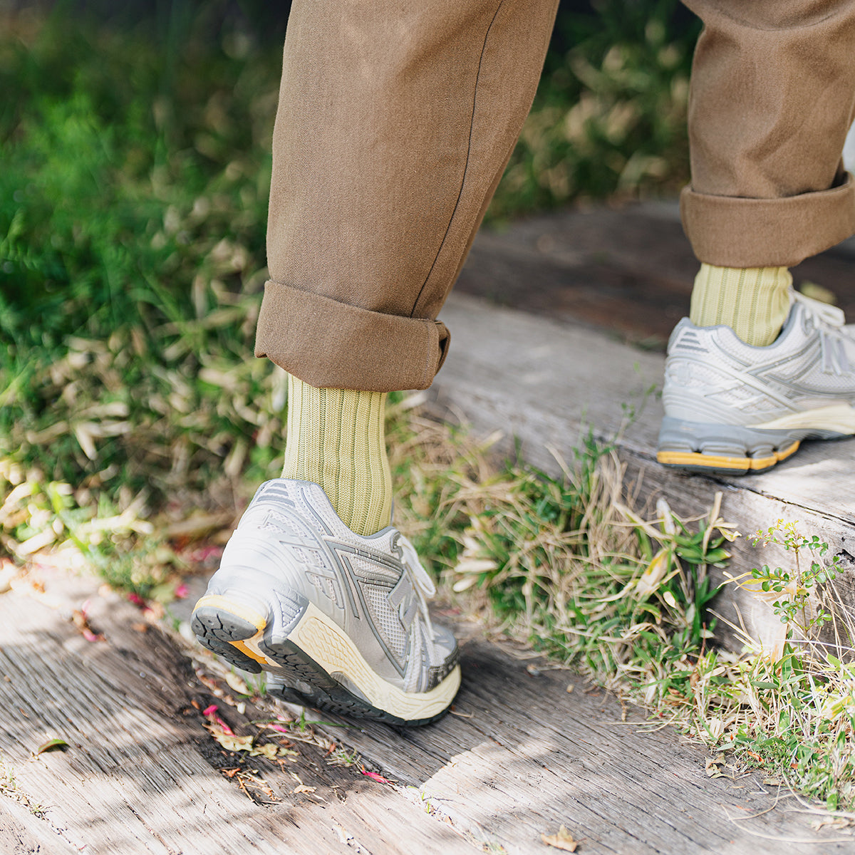 Person's legs wearing brown pants, light green socks, and grey athletic shoes, stepping on wooden beam stairs in grass