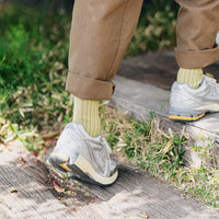 Person's legs wearing brown pants, light green socks, and grey athletic shoes, stepping on wooden beam stairs in grass