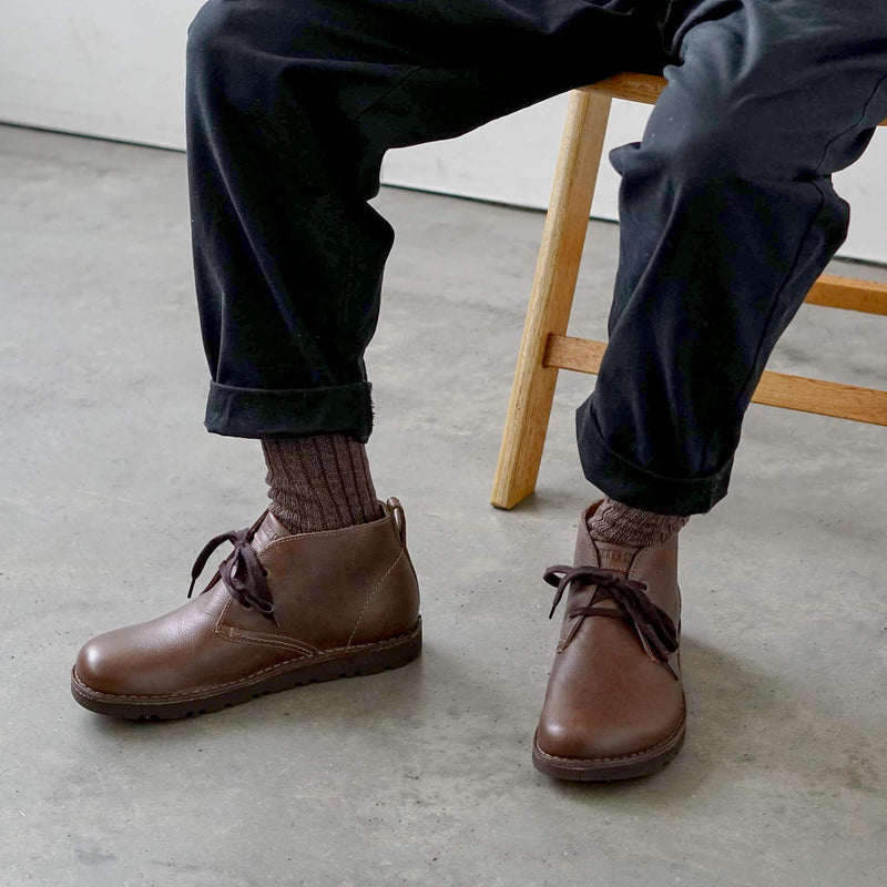 Person wearing brown leather shoes, socks, and dark pants sitting on a wooden stool.