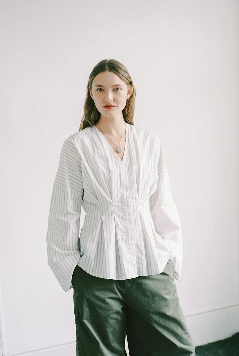 A woman wearing a white long-sleeve blouse with thin vertical gray stripes and a gathered waist, paired with dark olive green pants, standing against a plain white wall.
