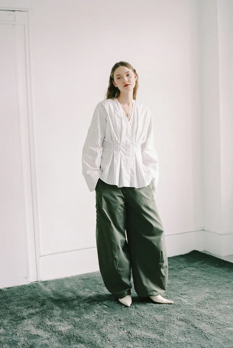 A woman stands in a minimalist white room wearing a white long-sleeve pleated blouse and dark olive green wide-leg trousers.