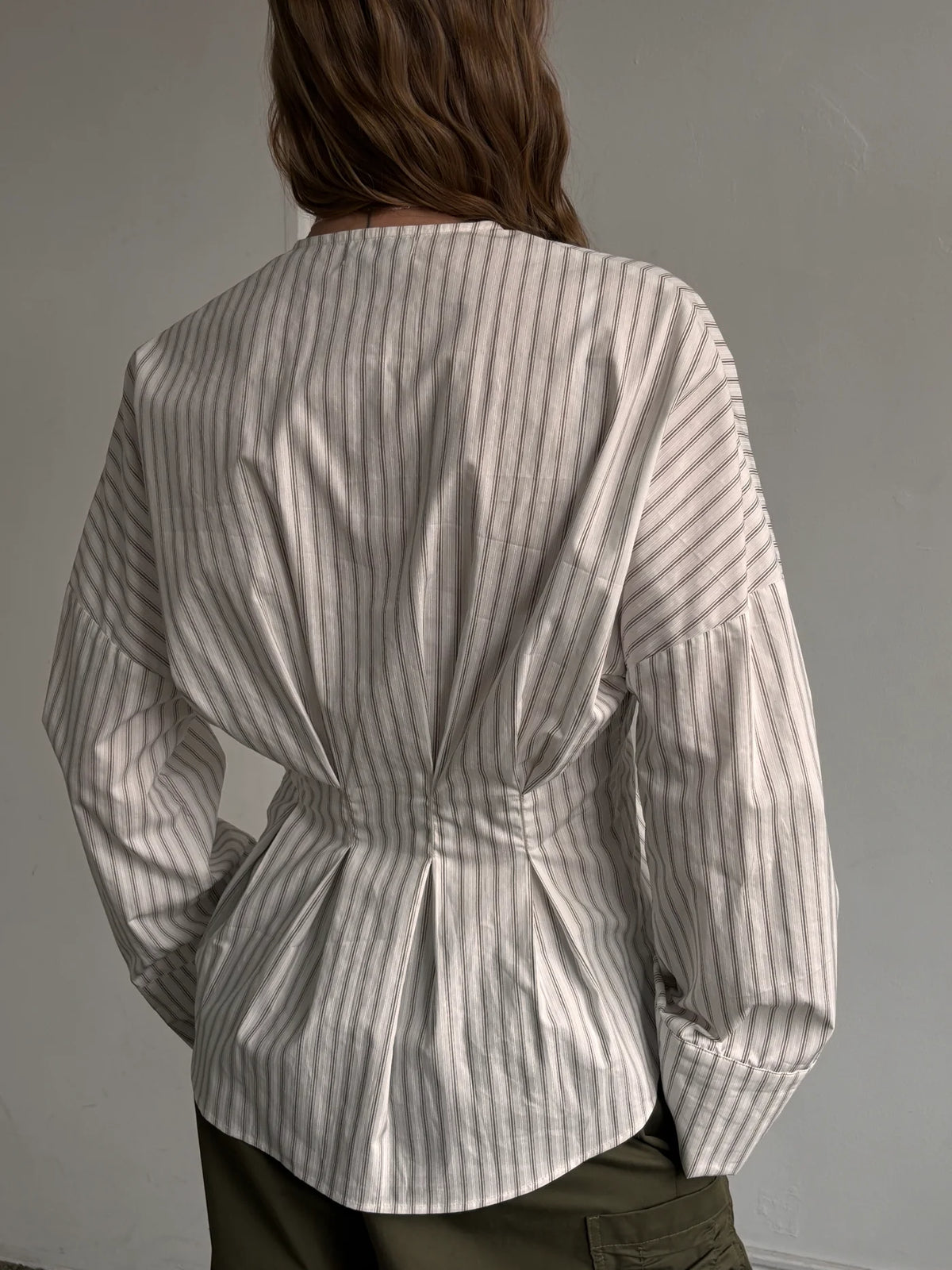 A person shown from the back wearing a long-sleeve striped blouse with gathered detailing at the waist and cuffs, paired with dark olive pants against a plain light gray wall.