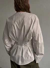 A person shown from the back wearing a long-sleeve striped blouse with gathered detailing at the waist and cuffs, paired with dark olive pants against a plain light gray wall.