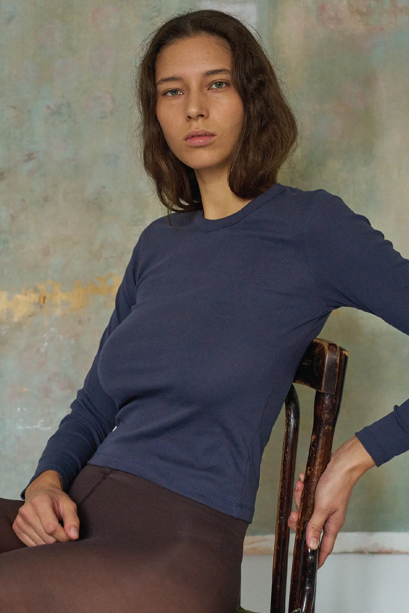 Woman wearing a blue long-sleeve shirt sitting on a chair against a textured wall.