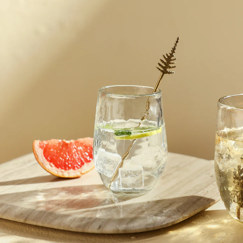 Pebbled drinking glasses with fruit water and a stir stick on a natural surface