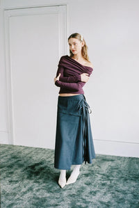 A woman stands in a minimalist room with a white wall and door, wearing a dark plum off-the-shoulder long-sleeve crop top and a dark teal wrap midi skirt with white pointed-toe heels.
