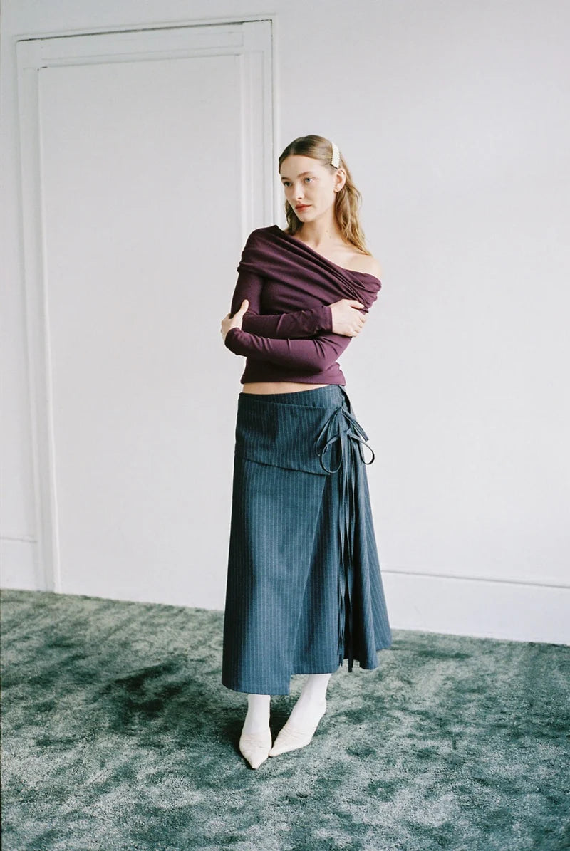 A woman stands in a minimalist room with a white wall and door, wearing a dark plum off-the-shoulder long-sleeve crop top and a dark teal wrap midi skirt with white pointed-toe heels.