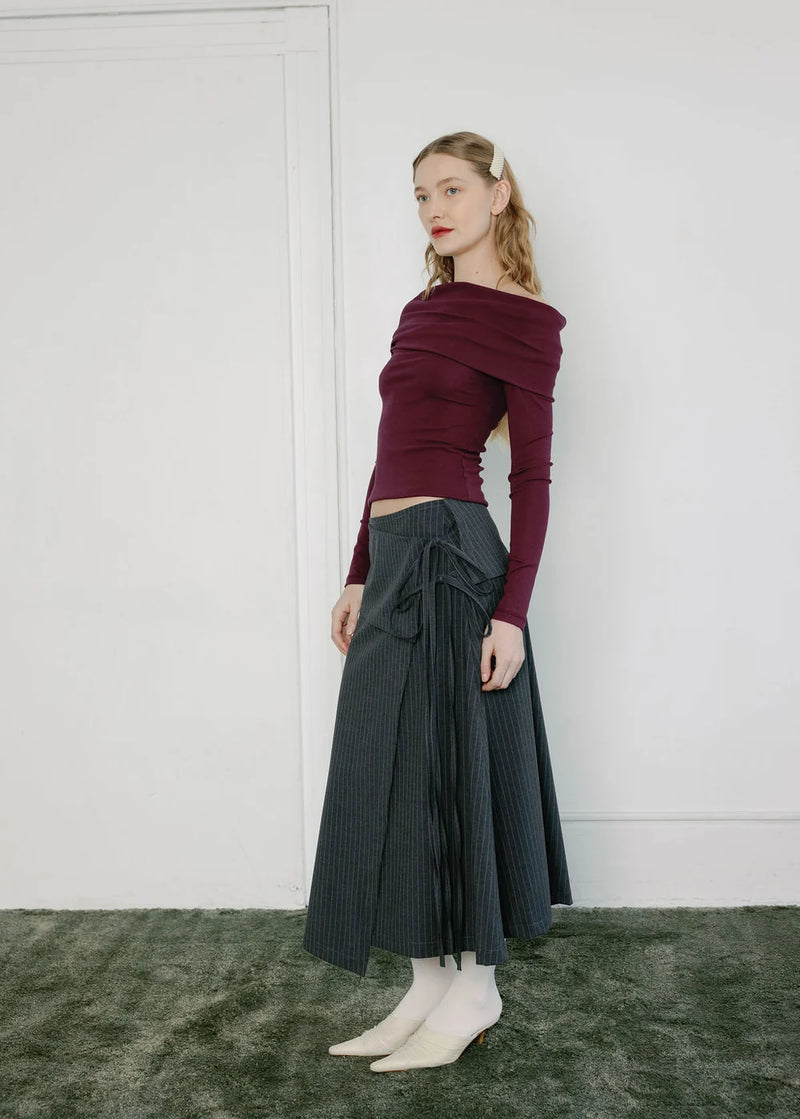 A woman stands in a minimalist room wearing a dark burgundy off-the-shoulder long-sleeve top and a dark gray pleated midi skirt with a side bow, paired with white pointed-toe flats.