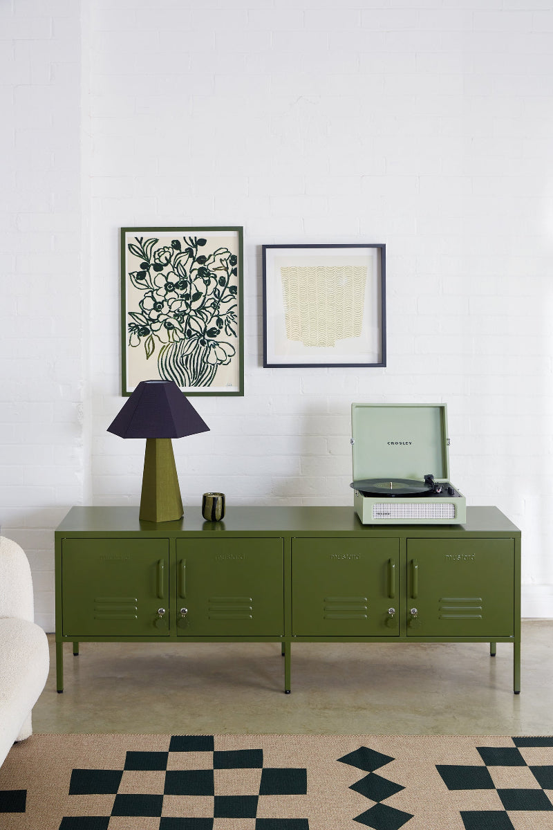 Green cabinet with a lamp and record player against a white wall with framed artwork.