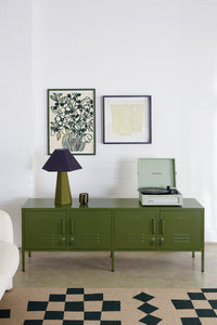 Green cabinet with a lamp and record player against a white wall with framed artwork.