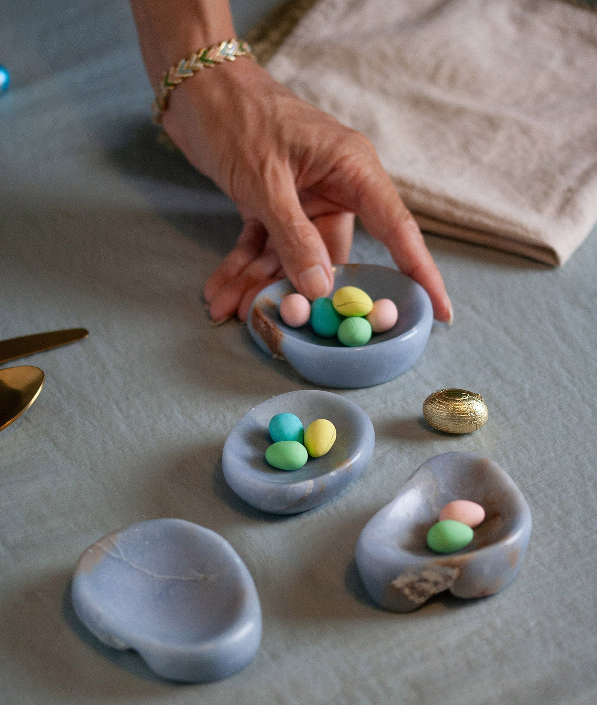 Four light blue stone dishes holding egg shaped candies on a textile surface with a hand reaching for one dish