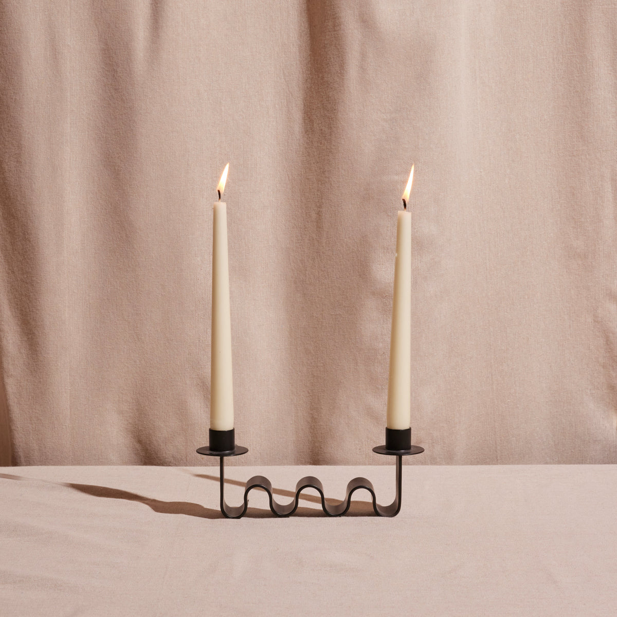 Black metal wavy candle holder with two lit candles against a beige curtain background