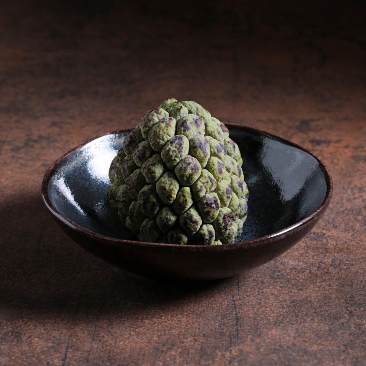 Green textured fruit like object in a dark ceramic bowl on a dark brown surface
