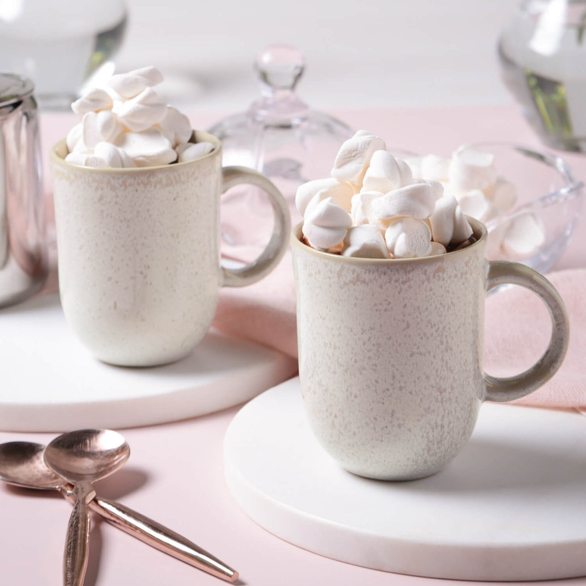 Two mugs filled with hot chocolate and marshmallows on a pink surface.