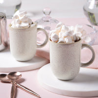 Two mugs filled with hot chocolate and marshmallows on a pink surface.