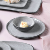 Gray ceramic dish with a small ramekin containing a piece of food on a pink surface.