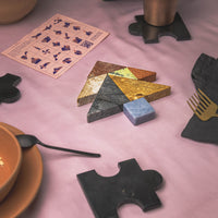 Colorful tangram puzzle pieces on a pink surface with a cup and utensils.