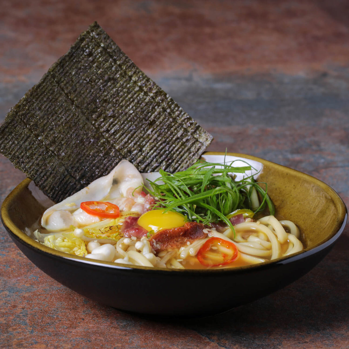 Bowl of udon noodles with vegetables and a piece of seaweed on a textured surface