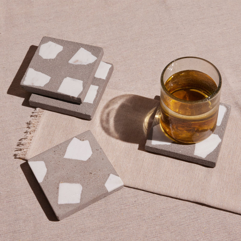 Set of square stone coasters with white patterns on a beige surface, one coaster holding a glass of amber liquid.