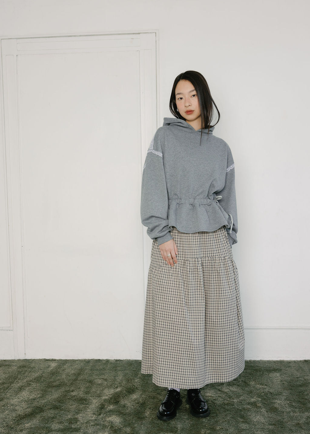 A model stands in a minimalist room wearing a gray hoodie with white sleeve lace detail and a beige-and-black gingham pleated maxi skirt, paired with black shoes.