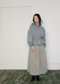 A model stands in a minimalist room wearing a gray hoodie with white sleeve lace detail and a beige-and-black gingham pleated maxi skirt, paired with black shoes.