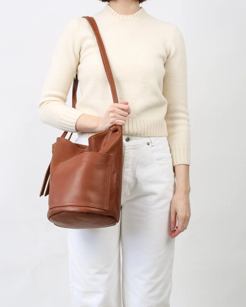A person wearing a cream knit sweater and white pants carries a brown leather bucket bag with a shoulder strap against a plain light background.