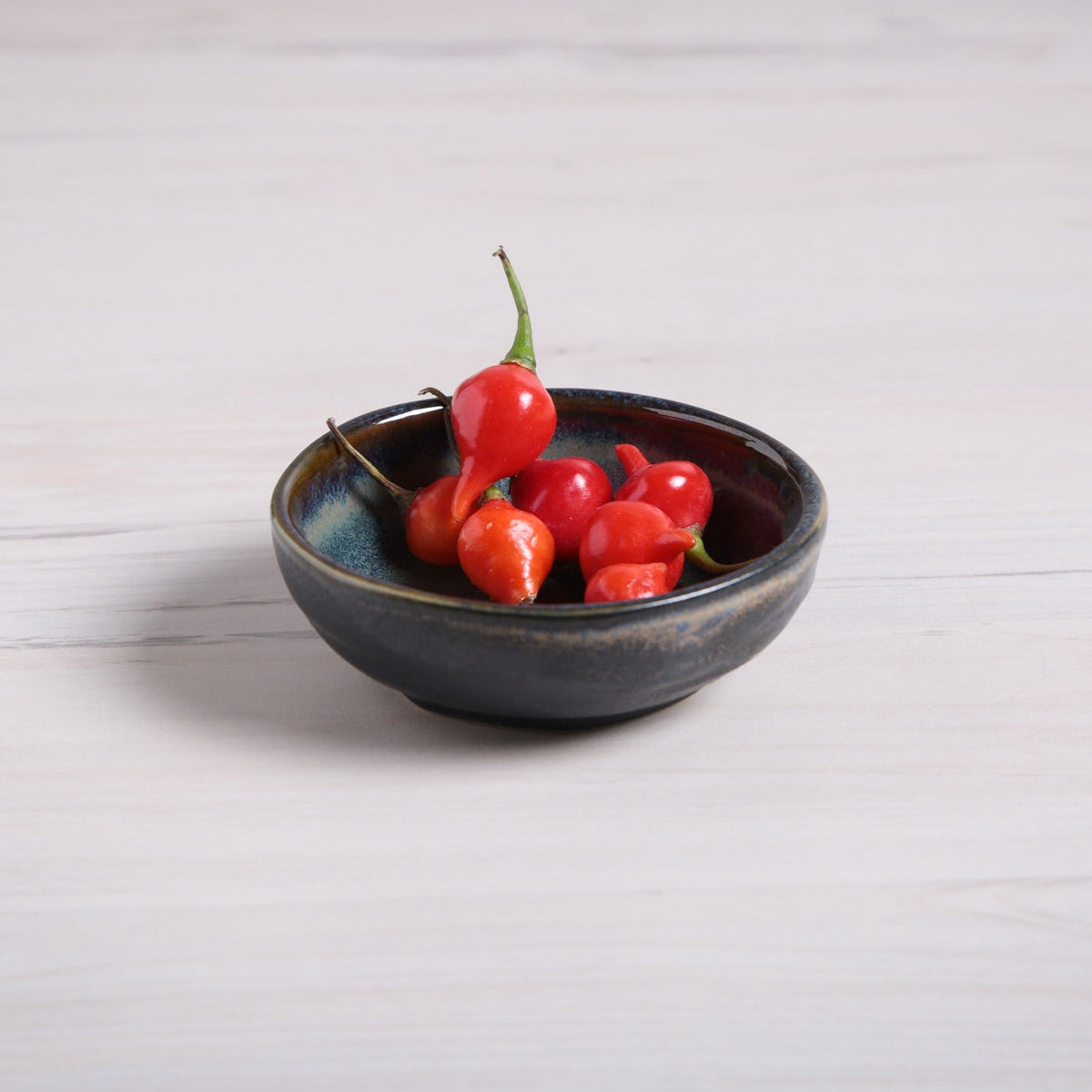 Black ceramic ramekin with red chili peppers on a light wooden surface