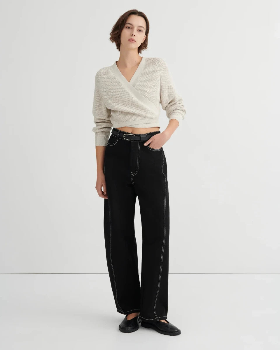 A model stands against a plain light gray studio background wearing a cream wrap-style knit sweater, black wide-leg jeans with contrast stitching, and black flats.