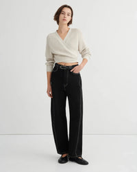 A model stands against a plain light gray studio background wearing a cream wrap-style knit sweater, black wide-leg jeans with contrast stitching, and black flats.