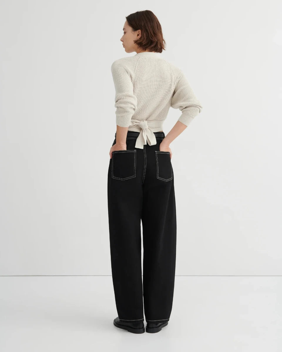 A woman stands with her back to the camera wearing a cream ribbed knit sweater tied at the waist and black wide-leg trousers with contrast stitching, against a plain white studio background.