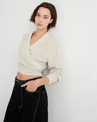 A woman wearing a cream-colored ribbed knit wrap cardigan and black pants, standing against a plain light gray wall.