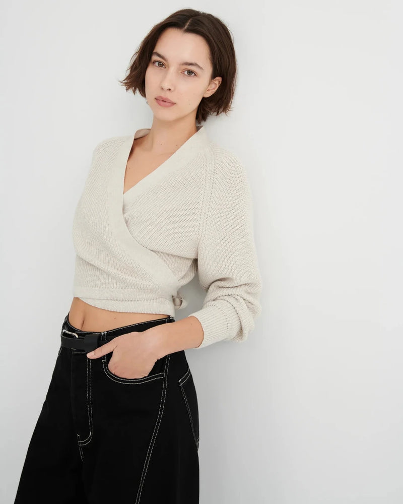 A woman wearing a cream-colored ribbed knit wrap cardigan and black pants, standing against a plain light gray wall.