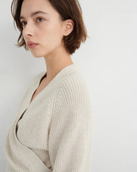 A woman with short brown hair wearing a cream-colored knit wrap cardigan, shown in a close-up profile against a light gray background.