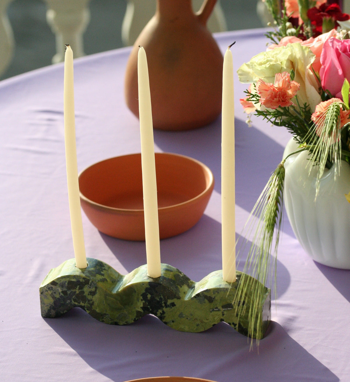 Decorative stone candle holder with taper candles on a table with a vase and flowers in the background.