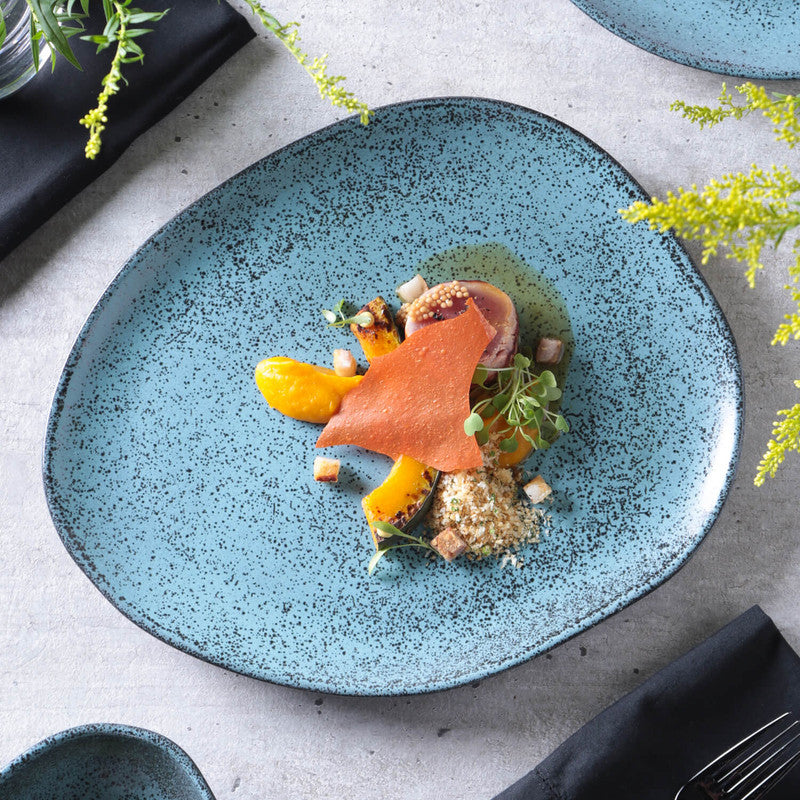 Blue speckled ceramic plate with a gourmet dish on a light gray surface with greenery.