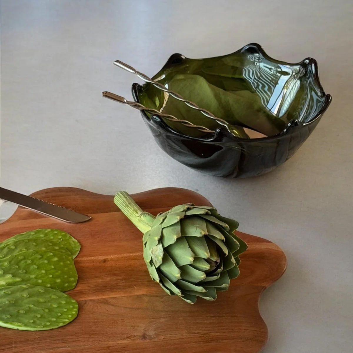 Green artichoke on a wooden cutting board with a decorative green glass bowl in the background.