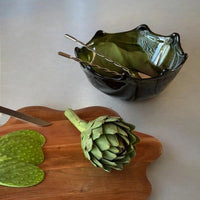 Green artichoke on a wooden cutting board with a decorative green glass bowl in the background.
