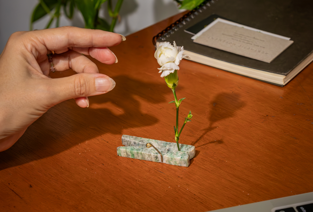 green stone clothespin holding a flower stem on a desk surface with a hand reaching toward it