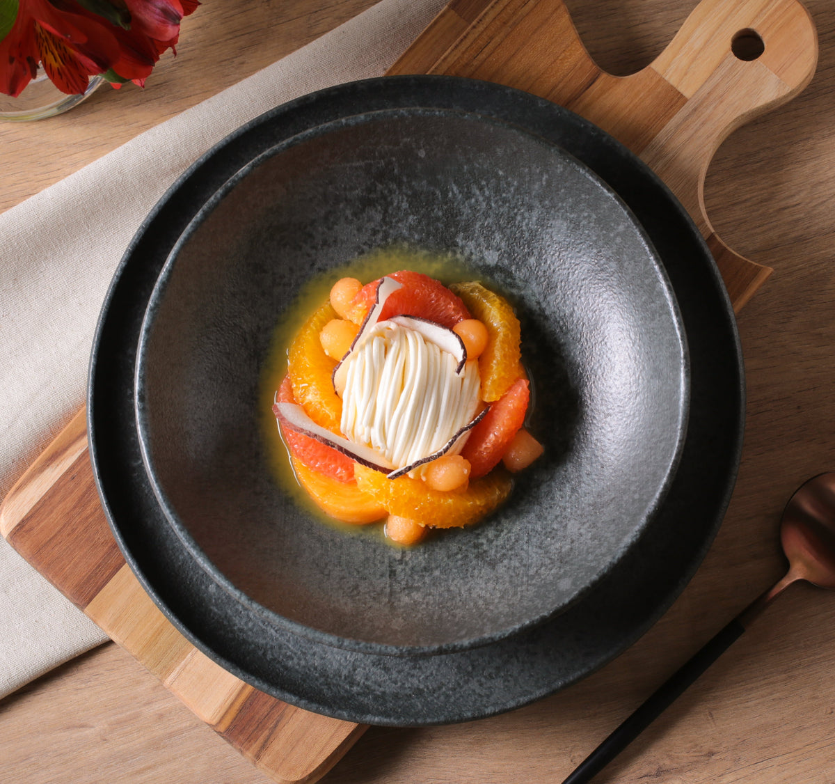 Delicate dessert presentation on a dark plate with a wooden cutting board and flowers in the background