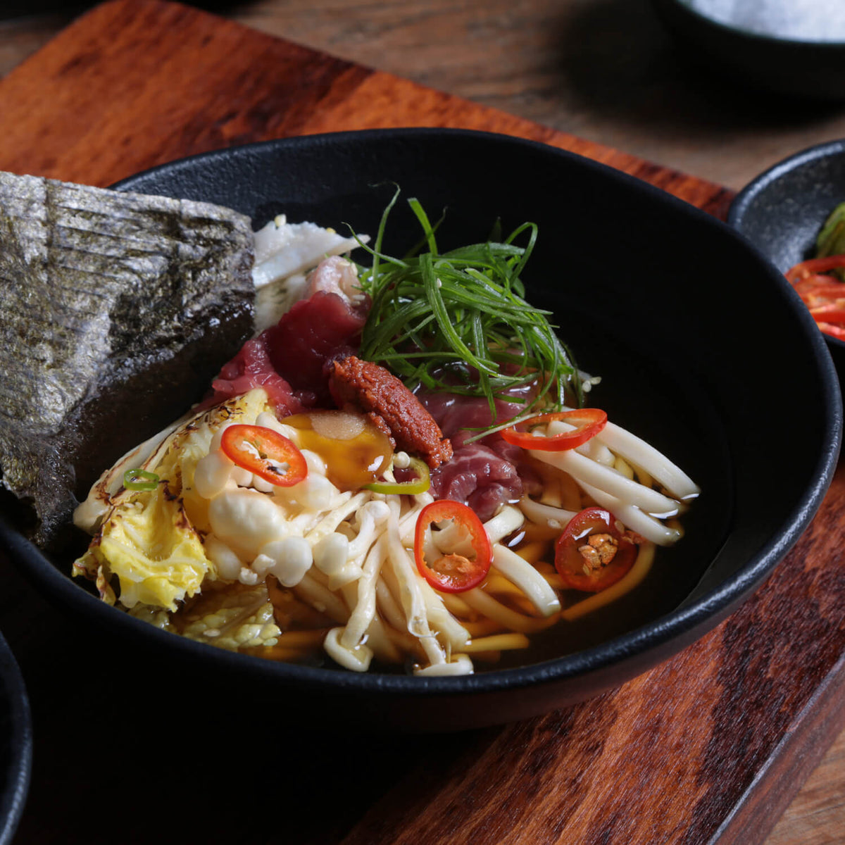 Noodle dish with vegetables and red chili peppers in a black bowl on a wooden surface